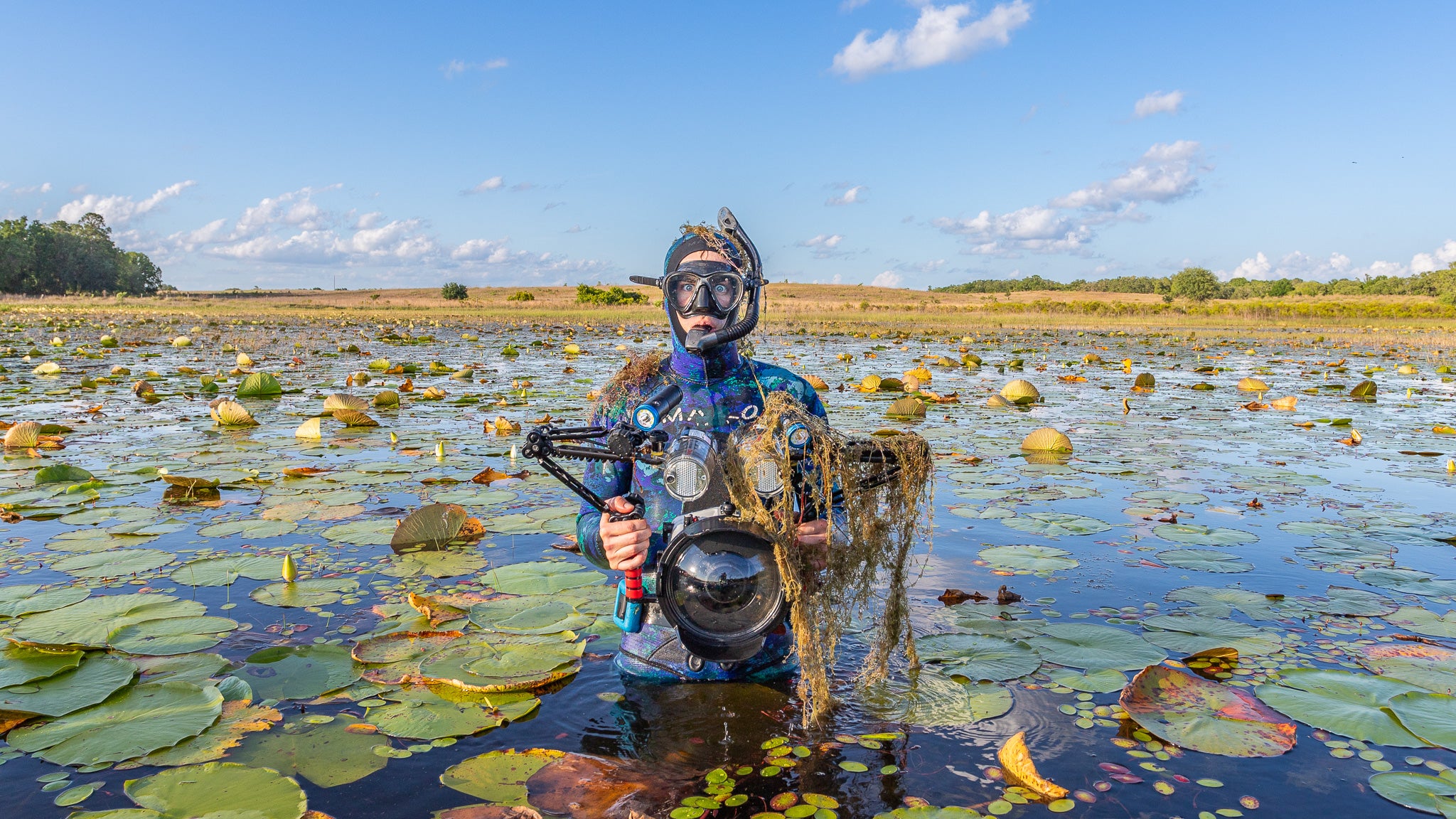 Person in snorkeling gear holding a underwater camera covered in pond weeds with lily pads and sky in the background